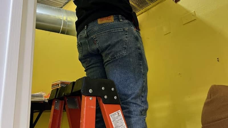 A Luxe Ducts technician on a ladder lifting a return air grille off a wall in a Chicago home